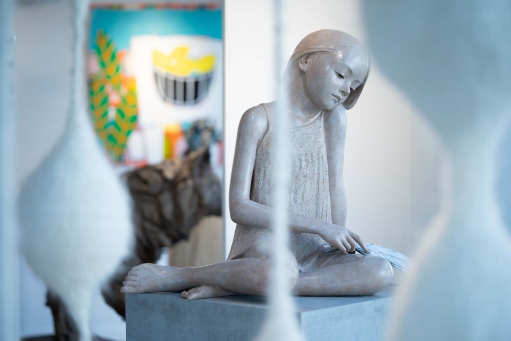 Oeuvre de l'artiste Berit, en bronze imitant la patine rose d'une pierre en granit. On aperçoit plus loin d'autres oeuvres, notamment des peintures aux contrastes saisissants exposés dans la galerie d'art.