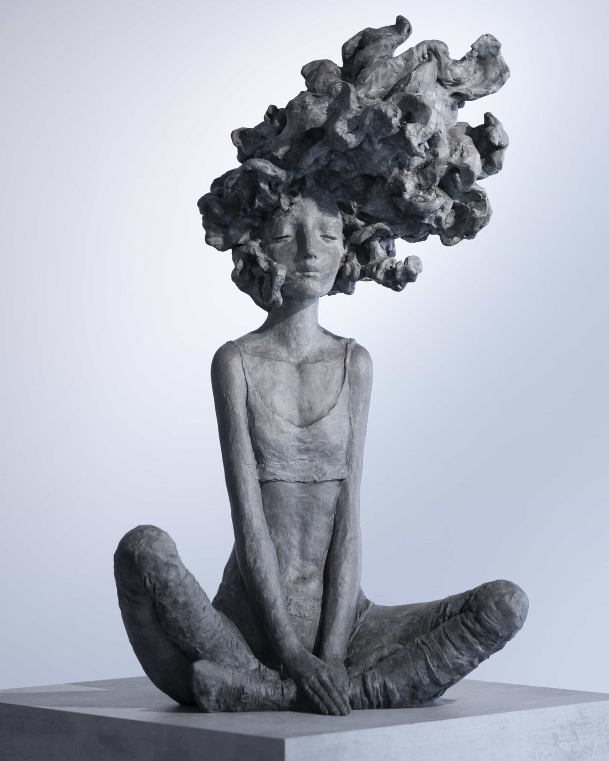 Valérie Hadida, dans *Nouvelle vague*, propose une sculpture figurative où l’intériorité psychique semble prendre forme matérielle. L’œuvre représente une jeune femme assise au sol, dans une posture calme et presque méditative. Les jambes ouvertes et repliées créent une assise stable, tandis que les bras allongés convergent vers les mains jointes, donnant à la figure une verticalité silencieuse. Le premier élément qui capte le regard est la chevelure monumentale, transformée en masse organique et tourbillonnante. Cette matière déborde largement les proportions naturelles du corps et agit comme une extension mentale ou émotionnelle du personnage. La “vague” du titre peut alors être comprise de plusieurs façons : * une vague intérieure, faite de pensées, de rêves ou d’émotions ; * une énergie créatrice en expansion ; * un mouvement naturel, proche du vent, de l’eau ou du nuage ; * ou encore une métaphore de transformation personnelle. La tension entre immobilité et mouvement structure toute l’œuvre. Le corps est calme, presque immobile, mais la tête semble traversée par un flux incessant. Cette opposition crée une sensation poétique : l’être est ancré physiquement, tandis que l’esprit dérive, s’élève ou se déploie. Le traitement de la matière participe fortement à cette impression. La surface rugueuse, modelée de manière expressive, conserve les traces du geste sculptural. On perçoit une matière vivante, vibrante, loin d’un rendu lisse ou académique. Cela donne à la sculpture une présence tactile et émotionnelle. La patine grise ou bronze sombre accentue également la dimension minérale et intemporelle de la figure. L’esthétique de Valérie Hadida se situe souvent entre figuration et imaginaire. Les corps sont allongés, fragiles, presque suspendus dans un état intérieur. Dans *Nouvelle vague*, cette fragilité corporelle contraste avec l’ampleur presque cosmique de la chevelure. Le personnage paraît à la fois vulnérable et habité d’une force invisible. L’œuvre peut évoquer plusieurs filiations : * certaines recherches expressionnistes autour de la matière ; * l’univers onirique du symbolisme ; * ou encore une approche contemporaine de la sculpture psychologique, où le corps devient le support d’états mentaux. Ce qui rend cette sculpture particulièrement forte, c’est sa capacité à rester ouverte à l’interprétation. Elle peut être lue comme : * une célébration de l’imagination ; * une représentation de la vie intérieure féminine ; * une métaphore du tumulte émotionnel ; * ou un moment de paix au cœur du chaos. ### En résumé *Nouvelle vague* associe : * une posture méditative, * une matière expressive, * et une chevelure transformée en phénomène organique et mental. Valérie Hadida y construit une figure à la fois intime et universelle, où l’intériorité devient paysage sculptural.