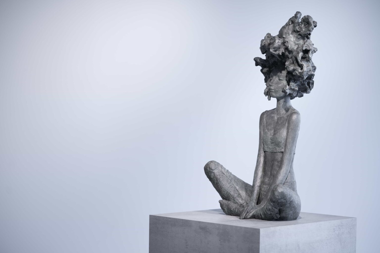 Valérie Hadida, dans *Nouvelle vague*, propose une sculpture figurative où l’intériorité psychique semble prendre forme matérielle. L’œuvre représente une jeune femme assise au sol, dans une posture calme et presque méditative. Les jambes ouvertes et repliées créent une assise stable, tandis que les bras allongés convergent vers les mains jointes, donnant à la figure une verticalité silencieuse. Le premier élément qui capte le regard est la chevelure monumentale, transformée en masse organique et tourbillonnante. Cette matière déborde largement les proportions naturelles du corps et agit comme une extension mentale ou émotionnelle du personnage. La “vague” du titre peut alors être comprise de plusieurs façons : * une vague intérieure, faite de pensées, de rêves ou d’émotions ; * une énergie créatrice en expansion ; * un mouvement naturel, proche du vent, de l’eau ou du nuage ; * ou encore une métaphore de transformation personnelle. La tension entre immobilité et mouvement structure toute l’œuvre. Le corps est calme, presque immobile, mais la tête semble traversée par un flux incessant. Cette opposition crée une sensation poétique : l’être est ancré physiquement, tandis que l’esprit dérive, s’élève ou se déploie. Le traitement de la matière participe fortement à cette impression. La surface rugueuse, modelée de manière expressive, conserve les traces du geste sculptural. On perçoit une matière vivante, vibrante, loin d’un rendu lisse ou académique. Cela donne à la sculpture une présence tactile et émotionnelle. La patine grise ou bronze sombre accentue également la dimension minérale et intemporelle de la figure. L’esthétique de Valérie Hadida se situe souvent entre figuration et imaginaire. Les corps sont allongés, fragiles, presque suspendus dans un état intérieur. Dans *Nouvelle vague*, cette fragilité corporelle contraste avec l’ampleur presque cosmique de la chevelure. Le personnage paraît à la fois vulnérable et habité d’une force invisible. L’œuvre peut évoquer plusieurs filiations : * certaines recherches expressionnistes autour de la matière ; * l’univers onirique du symbolisme ; * ou encore une approche contemporaine de la sculpture psychologique, où le corps devient le support d’états mentaux. Ce qui rend cette sculpture particulièrement forte, c’est sa capacité à rester ouverte à l’interprétation. Elle peut être lue comme : * une célébration de l’imagination ; * une représentation de la vie intérieure féminine ; * une métaphore du tumulte émotionnel ; * ou un moment de paix au cœur du chaos. ### En résumé *Nouvelle vague* associe : * une posture méditative, * une matière expressive, * et une chevelure transformée en phénomène organique et mental. Valérie Hadida y construit une figure à la fois intime et universelle, où l’intériorité devient paysage sculptural.