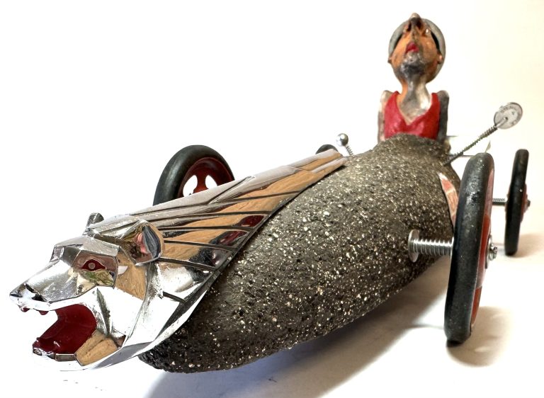 L’image présente une sculpture-objet au caractère décalé, mêlant matériaux bruts, pièces métalliques et une figure humaine au style expressif. Voici une description détaillée : Cette œuvre représente un véhicule imaginaire construit autour d’un corps massif, granuleux et sombre, rappelant la texture de la pierre ou du béton. La forme générale évoque un bolide allongé et aérodynamique. À l’avant, un imposant élément métallique chromé sculpté en tête de lion, gueule ouverte, confère toute sa puissance visuelle à la pièce. Son style anguleux et brillant contraste fortement avec la matière rugueuse du châssis, créant un dialogue entre noblesse mécanique et rusticité. Les roues, fixées par de longues vis apparentes, renforcent l’aspect bricolé et inventif typique d’une esthétique de récupération. Elles présentent un mélange de noir et de rouge, ajoutant une touche dynamique à la structure. À l’arrière du véhicule, un personnage sculpté émerge du corps de la machine. Son torse peint, visiblement doté d’un vêtement rouge, et son visage expressif, la tête rejetée en arrière, suggèrent une posture dramatique ou exaltée. Les traits accentués, faits de volumes irréguliers et de couleurs franches, lui donnent une allure presque théâtrale. Ce pilote improbable contribue grandement à l’humour et au caractère narratif de la sculpture. L’ensemble combine ainsi matières industrielles, modelage manuel, récupérations d’objets et imagination débridée. Le résultat est un véhicule-poème, à la fois humoristique, brut et précieux, emblématique d’une démarche artistique qui transforme les rebuts en personnages et machines porteuses d’histoire. **Résumé actionnable :** L’œuvre mêle un châssis granuleux, une tête de lion chromée à l’avant, des roues bricolées et un personnage expressif à l’arrière, créant un bolide sculptural hybride entre fantaisie, récupération et narration visuelle.
