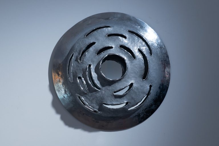 Œuvre murale circulaire en aluminium poli, Ether n°2 de Kymia se présente comme un miroir-sculpture où la surface métallique est perforée de fentes courbes disposées en vortex autour d’un noyau central évidé, suggérant le mouvement d’un tourbillon d’énergie immatérielle. [ppl-ai-file-upload.s3.amazonaws](https://ppl-ai-file-upload.s3.amazonaws.com/web/direct-files/attachments/images/89356762/68111593-6195-4e0f-8dd4-a482acfe5955/KYMIA-Ether-ndeg2-Aluminium-D33cm-2.jpg?AWSAccessKeyId=ASIA2F3EMEYE3BRQA6OI&Signature=eF445ui%2BgGds8y8fvhi7jGvgNmw%3D&x-amz-security-token=IQoJb3JpZ2luX2VjEC8aCXVzLWVhc3QtMSJGMEQCIHr5aGj%2BuGr7YL8yE%2Fs5T1LqmBlXk5Haftumv0SBGz8qAiBhD9GP%2BYq0EKzPDYvMzs1kv%2FC863nHXWVt9Rngt6XOwir8BAj4%2F%2F%2F%2F%2F%2F%2F%2F%2F%2F8BEAEaDDY5OTc1MzMwOTcwNSIMLV0IjAhhSRCbU0V6KtAEqSYm%2FLhZQjKB52Z9RxG%2F69D9jE6SLS7DNeAAdK%2F1CfvYWmgfWTNJZv4KQi1qTjo2OoCvmujyYHyy%2Fk62aLPzpAVFCsIXB1RUWUdH%2BngbLANDemsrJ5PxniDifMhmnH84PF65sesDr4Uh1ibcpZFZ5WAuNTgV12vVOTIlPnMusm%2BObdr2%2F1u1GOIFy%2BBBN4xz6iFzLMasXFU5bkRanJ0szOiov5IQ92pNgre87L%2FIDeDAZE6JmhrFEbb2Krk%2B5zu%2FguqQruisLdB76H2Q9N1it7%2Bpr85uWlzlHo1AkuiUzct1s%2FB69HF5blTkmQljSz0rvPqIIqNsdPz1ZYTvZ%2FaRcYmYAeIV4lv6kQ0Rd%2FP8DCQzFI0YqXzv9H9xnfKTM2ejpNsFBuC5d19%2BoD3WGF8K%2B1ANO1KRvUNT9WkCkNJ9hzA6iBkEDxoYAVVkg8%2F1Mk9vVHyiJfhnI0%2F1%2BSE8R7JVAxhGdywVDui%2BH0Ph%2FIGhp1Nw%2BGcnwn5oZB3tZTzkjeEN8L8F140cGBITtMTsDFhwVClrQw5EhdOnebxIO7SRYIj8DxfcNMLuhT%2B33qDqNtK%2FyUca7WYxBIl4V6pq9Q4DquGPY%2BBO2CXxDIhoOMhPJzAZel5f4AFjDOo%2F0YHsPgH1pVgYonVey7DmBwKILeY6yCTHmY55HUO12fZKvhKX2zLuRpXhFBE8qgIO2QWxQfuQlLhMEGqqpd2ASCB2ikCB4L%2FNSCcR3mrr3qP8S%2ByogAlF%2BSNHQ5LGilyq9%2BxqCT5T1SmW3MOMOWB861b7V2LspzD2ic7LBjqZAYK6P0iplqVXTGlhHYqRTqaLHRZwzJxWDsjaZi6Xku06py7qonmlnDa4v4Vtd3W70YiO1%2F75u4xf5Oc5zvlbag2ZOAXy2mwkFlj0skE0ge4UJaOJn2Y5Trk2r01XYlySmmPCeK2WdpMdnidKbapU6pITOm0S9kmfLD00BFQSdfTWDEwv6ZNoye4JKTNzC1eEJnXwAhYEZUXXEA%3D%3D&Expires=1769180152) ## Forme et matière - L’œuvre adopte un disque bombé d’environ 33 cm de diamètre, entièrement façonné en **aluminium**, dont la peau martelée et polie joue avec les reflets de la lumière comme un miroir organique. [galerie-platini](https://www.galerie-platini.com/oeuvres/miroir-mobilier-art/) - Les découpes allongées et irrégulières, orientées en spirale, laissent passer le vide et créent des ombres mouvantes sur le mur, renforçant l’idée de flux et de circulation d’air ou d’éther. [ppl-ai-file-upload.s3.amazonaws](https://ppl-ai-file-upload.s3.amazonaws.com/web/direct-files/attachments/images/89356762/68111593-6195-4e0f-8dd4-a482acfe5955/KYMIA-Ether-ndeg2-Aluminium-D33cm-2.jpg?AWSAccessKeyId=ASIA2F3EMEYE3BRQA6OI&Signature=eF445ui%2BgGds8y8fvhi7jGvgNmw%3D&x-amz-security-token=IQoJb3JpZ2luX2VjEC8aCXVzLWVhc3QtMSJGMEQCIHr5aGj%2BuGr7YL8yE%2Fs5T1LqmBlXk5Haftumv0SBGz8qAiBhD9GP%2BYq0EKzPDYvMzs1kv%2FC863nHXWVt9Rngt6XOwir8BAj4%2F%2F%2F%2F%2F%2F%2F%2F%2F%2F8BEAEaDDY5OTc1MzMwOTcwNSIMLV0IjAhhSRCbU0V6KtAEqSYm%2FLhZQjKB52Z9RxG%2F69D9jE6SLS7DNeAAdK%2F1CfvYWmgfWTNJZv4KQi1qTjo2OoCvmujyYHyy%2Fk62aLPzpAVFCsIXB1RUWUdH%2BngbLANDemsrJ5PxniDifMhmnH84PF65sesDr4Uh1ibcpZFZ5WAuNTgV12vVOTIlPnMusm%2BObdr2%2F1u1GOIFy%2BBBN4xz6iFzLMasXFU5bkRanJ0szOiov5IQ92pNgre87L%2FIDeDAZE6JmhrFEbb2Krk%2B5zu%2FguqQruisLdB76H2Q9N1it7%2Bpr85uWlzlHo1AkuiUzct1s%2FB69HF5blTkmQljSz0rvPqIIqNsdPz1ZYTvZ%2FaRcYmYAeIV4lv6kQ0Rd%2FP8DCQzFI0YqXzv9H9xnfKTM2ejpNsFBuC5d19%2BoD3WGF8K%2B1ANO1KRvUNT9WkCkNJ9hzA6iBkEDxoYAVVkg8%2F1Mk9vVHyiJfhnI0%2F1%2BSE8R7JVAxhGdywVDui%2BH0Ph%2FIGhp1Nw%2BGcnwn5oZB3tZTzkjeEN8L8F140cGBITtMTsDFhwVClrQw5EhdOnebxIO7SRYIj8DxfcNMLuhT%2B33qDqNtK%2FyUca7WYxBIl4V6pq9Q4DquGPY%2BBO2CXxDIhoOMhPJzAZel5f4AFjDOo%2F0YHsPgH1pVgYonVey7DmBwKILeY6yCTHmY55HUO12fZKvhKX2zLuRpXhFBE8qgIO2QWxQfuQlLhMEGqqpd2ASCB2ikCB4L%2FNSCcR3mrr3qP8S%2ByogAlF%2BSNHQ5LGilyq9%2BxqCT5T1SmW3MOMOWB861b7V2LspzD2ic7LBjqZAYK6P0iplqVXTGlhHYqRTqaLHRZwzJxWDsjaZi6Xku06py7qonmlnDa4v4Vtd3W70YiO1%2F75u4xf5Oc5zvlbag2ZOAXy2mwkFlj0skE0ge4UJaOJn2Y5Trk2r01XYlySmmPCeK2WdpMdnidKbapU6pITOm0S9kmfLD00BFQSdfTWDEwv6ZNoye4JKTNzC1eEJnXwAhYEZUXXEA%3D%3D&Expires=1769180152) ## Sens et symbolique - Le vortex de fentes autour du centre vide évoque l’éther comme une énergie subtile, invisible mais structurante, qui relie le plein et le vide, le matériel et l’immatériel. [galerie-platini](https://www.galerie-platini.com/oeuvres/miroir-mobilier-art/) - En jouant sur la réflexion du spectateur dans le métal, l’œuvre fonctionne comme un miroir introspectif qui interroge la place du corps dans cet espace énergétique et cosmique.