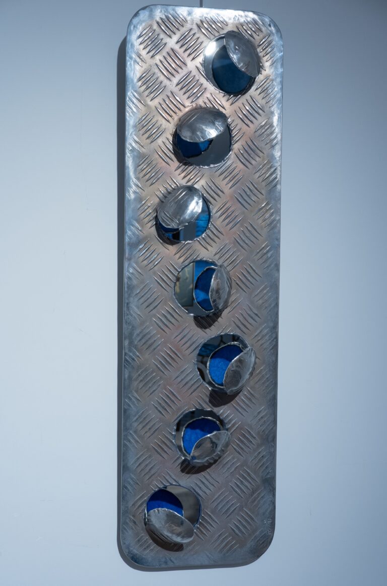 Cette œuvre murale de **Kymia** s’inscrit pleinement dans la démarche singulière de l’artiste, fondée sur une exploration sensible et instinctive du métal. Réalisée en aluminium martelé, elle se présente comme une plaque verticale perforée de cavités circulaires dont les rebords semblent arrachés, tordus ou entrouverts, laissant apparaître un fond bleu profond. Ce contraste entre la froideur industrielle du matériau et la vibration colorée crée une tension visuelle immédiate. Formée aux techniques de la chaudronnerie et du travail de l’aluminium après un long apprentissage en sculpture, Kymia développe une relation presque organique avec la matière, qu’elle ne cherche pas à dominer mais à accompagner dans ses réactions propres ([Galerie 21][1]). Ici, les déformations du métal évoquent des impacts, des éclosions ou des ouvertures, comme si la surface était traversée par une énergie interne. Le geste reste volontairement brut, laissant visibles les traces du feu, du martelage et de la torsion, caractéristiques de son travail. L’introduction du bleu — couleur souvent associée chez elle à une dimension intérieure ou spirituelle — transforme ces perforations en seuils visuels, invitant le regard à dépasser la surface pour accéder à une profondeur symbolique ([Galerie 21][1]). L’œuvre oscille ainsi entre abstraction et suggestion corporelle ou cosmique : autant de « blessures » que de « passages », entre matière et immatériel. Enfin, cette pièce reflète une constante dans son œuvre : détourner des références industrielles ou domestiques pour en faire des objets poétiques, où la violence du geste se conjugue à une recherche d’équilibre et de sens. L’aluminium, matériau central dans sa pratique, devient ici le vecteur d’une tension maîtrisée entre chaos et composition. **Résumé actionnable :** Œuvre en aluminium caractéristique de Kymia : travail direct de la matière, tension entre brutalité et poésie, jeu de perforations révélant une profondeur colorée et symbolique. [1]: https://galerie21.fr/artistes/kymia/?utm_source=chatgpt.com "Kymia | Galerie 21"