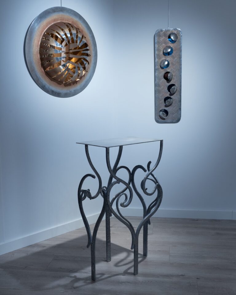 Cet ensemble présenté par **Kymia** met en évidence la cohérence de son langage plastique, tout en révélant la diversité de ses explorations formelles autour du métal. À travers trois pièces distinctes — une sculpture murale circulaire, un panneau perforé et une console aux lignes sinueuses — l’artiste développe un dialogue entre tension industrielle et élan organique. La pièce murale ronde, structurée comme une turbine ou une corolle métallique, déploie un mouvement centrifuge. Les lamelles découpées et repliées vers l’intérieur créent un effet de vortex, accentué par des tonalités chaudes qui contrastent avec la froideur périphérique du métal. Cette dynamique évoque une énergie contenue, presque mécanique, mais transposée dans un registre sensible. À ses côtés, le panneau vertical en aluminium martelé — déjà emblématique de sa pratique — introduit une dimension plus brute. Les perforations irrégulières, aux bords relevés, laissent apparaître une profondeur colorée, suggérant des ouvertures ou des impacts. Ce jeu entre surface fermée et percée intérieure traduit une réflexion récurrente chez Kymia sur la matière comme enveloppe vivante, susceptible d’être traversée ou transformée. Enfin, la console en métal forgé prolonge cette logique dans un registre fonctionnel détourné. Ses pieds, constitués de volutes élancées et entrelacées, rappellent des tiges végétales ou des arabesques en croissance. Le plateau, sobre et géométrique, vient stabiliser cet ensemble mouvant, créant un contraste entre rigueur structurelle et liberté formelle. Fidèle à sa formation technique, l’artiste maîtrise ici pleinement les contraintes de l’équilibre et de la solidité, tout en conservant une écriture gestuelle. Dans cet accrochage, Kymia confirme son approche intuitive du métal : un matériau qu’elle plie, découpe et assemble sans chercher à en effacer les tensions, mais au contraire à les révéler. Chaque œuvre oscille entre objet, sculpture et fragment d’organisme, participant à une esthétique de la transformation où le métal devient presque vivant. **Résumé actionnable :** Ensemble cohérent mêlant mural et mobilier : tension entre industriel et organique, travail du métal comme matière vivante, équilibre entre fonction, mouvement et expressivité.