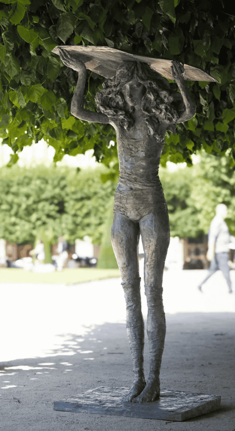 Sculpture en bronze représentant une figure humaine élancée portant une large forme plate au-dessus de sa tête, présentée en plein air sous un arbre, sur un socle bas.