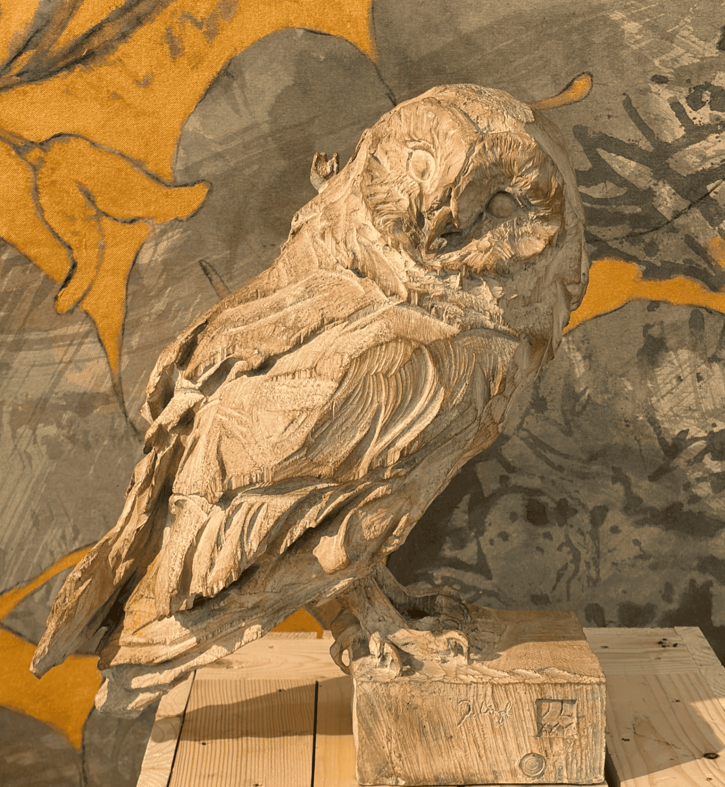 **Texte alternatif :** Sculpture en bois représentant un oiseau de proie aux ailes repliées, posé sur une caisse en bois, devant un décor peint de formes animales et végétales dans des tons ocres et gris.